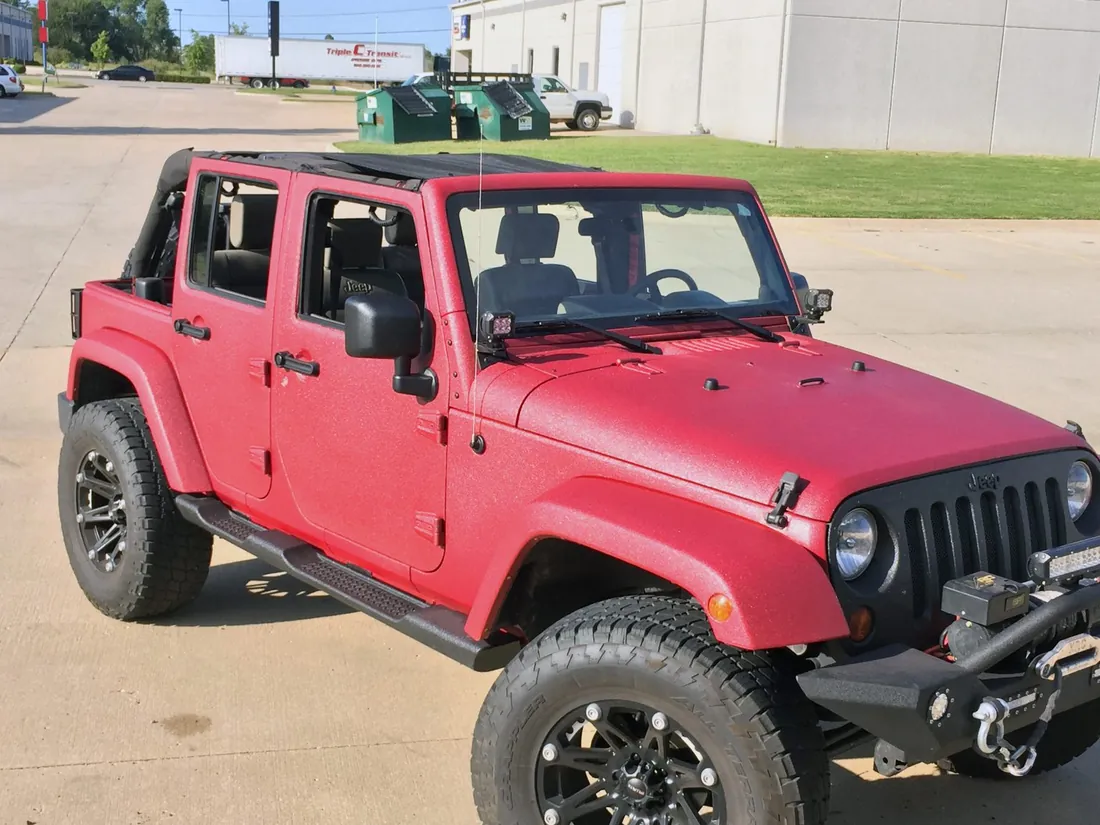 A Jeep Wrangler Unlimited coated head to toe in a metallic red textured protective coating, parked outside a shop.