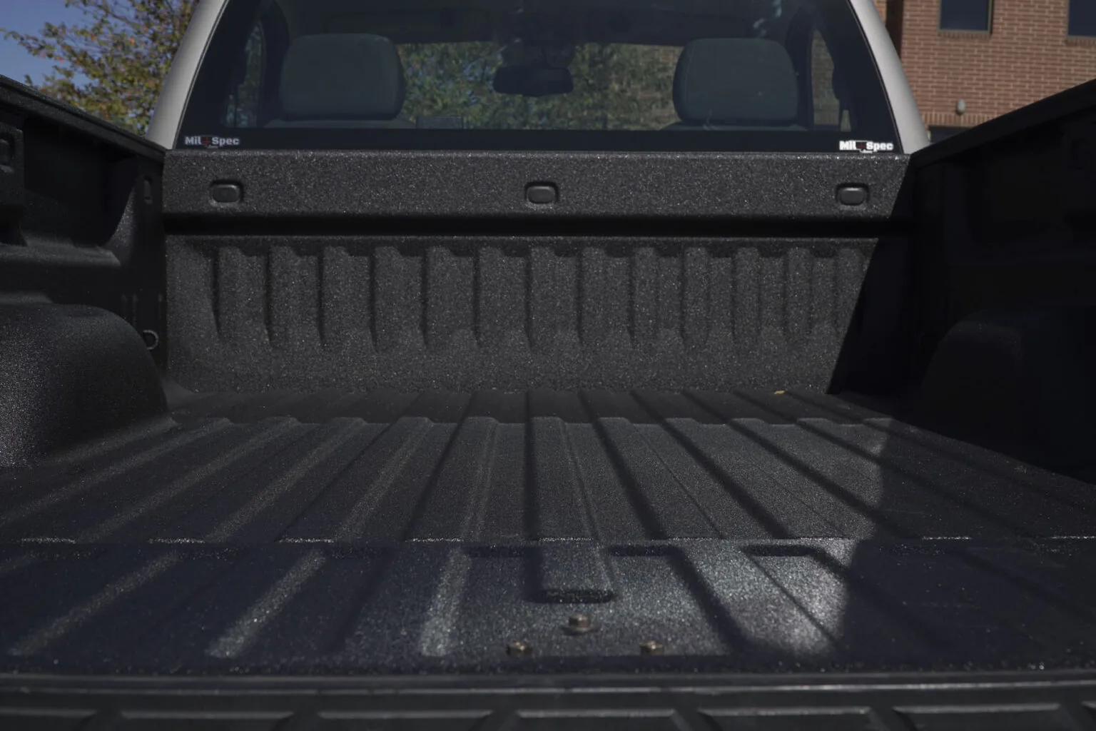 A Chevrolet pickup bed sprayed with a black textured Mil-Spec liner, seen from the open tailgate looking toward the cab, with the sprayed logo visible on the bulkhead.